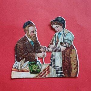 Op de afbeelding: Vintage papieren uitgesneden illustratie met twee figuren in traditionele Joodse kleding. Eén figuur, met baard en hoed, helpt de ander met het omwikkelen van hun arm. Een groene tas met een ster staat op een tafel.