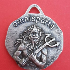 May include: A silver-toned metal pendant with a raised image of a bearded man holding a trident. The pendant is inscribed with the words "omnisports" and "succes".