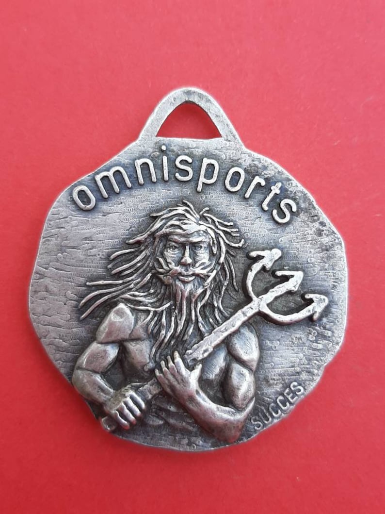 May include: A silver-toned metal pendant with a raised image of a bearded man holding a trident. The pendant is inscribed with the words "omnisports" and "succes".