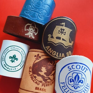 May include: Five leather patches with different designs. The patches are white, brown, blue, and black. The designs include a fleur-de-lis, a ship, a fish, and a scout logo. The text on the patches includes "Anglia 88", "Scout Fellowship", "Shia Ismaili Scouts Group", "G.E. Falcao Peregrino Brasil", and "Anhadadar".