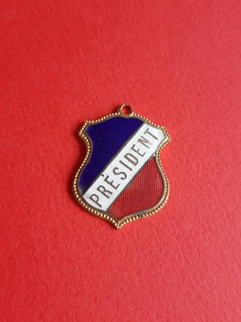 May include: A gold-colored metal shield-shaped charm with a blue, white, and red enamel design. The word "PRESIDENT" is written in white on the charm.