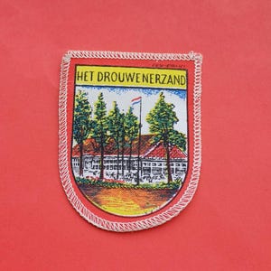Op de afbeelding: Een vintage geborduurde patch met een witte rand en een rood, geel en groen beeld van een gebouw met bomen en een vlag. De tekst "HET DROUWENERSAND" is in zwart op een witte achtergrond gedrukt.