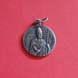 Op de afbeelding: Een zilverkleurige hanger met een gedetailleerde afbeelding van een religieuze figuur die een staf met een kruis vasthoudt. De hanger heeft de inscriptie "S. CORNELIUS" en "O.P.N." langs de rand. De achtergrond is effen rood.