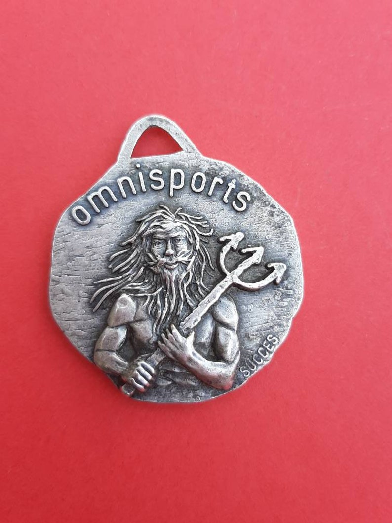May include: Silver-toned metal keychain with a raised image of a man holding a trident. The keychain is round and has the word "omnisports" written around the top edge. The word "succes" is written on the bottom edge.