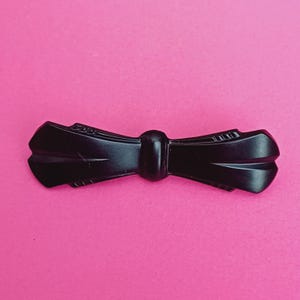 Op de afbeelding: Een zwarte haarclip in de vorm van een strik, met een afgeronde midden en gebogen uiteinden. De clip is gemaakt van een glanzend materiaal en staat tegen een felroze achtergrond. Het ontwerp is symmetrisch en heeft een vintage uitstraling.