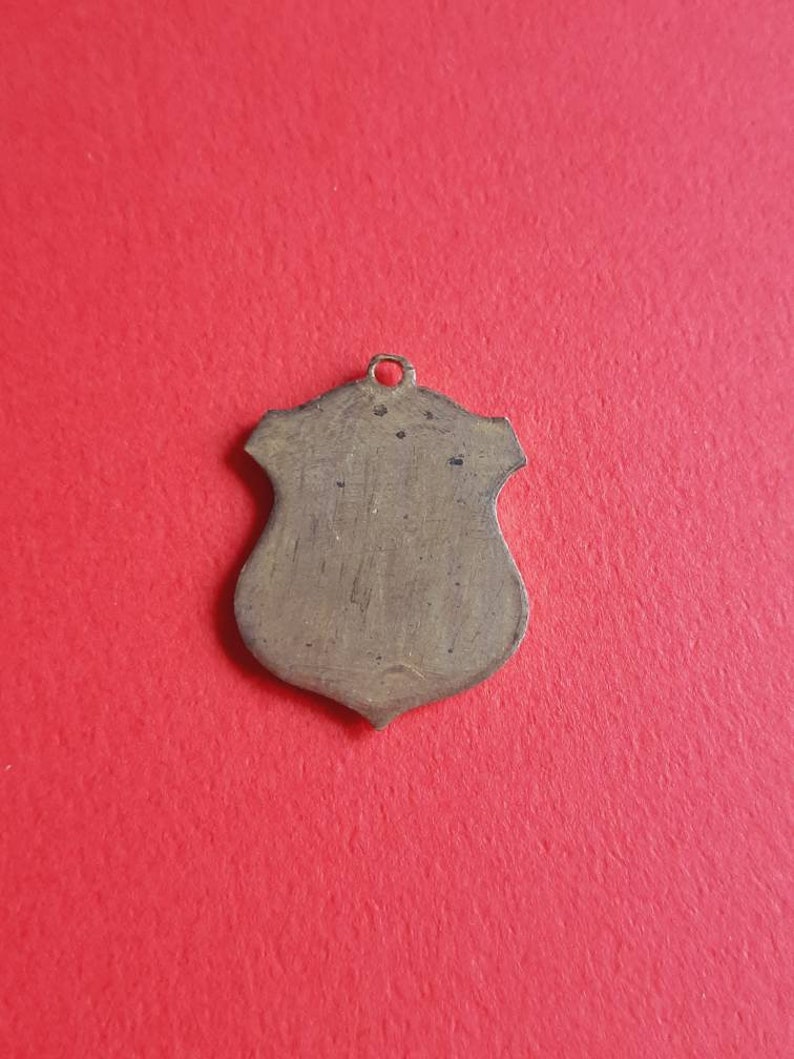 May include: A blank, gold-toned, shield-shaped pendant with a loop at the top.