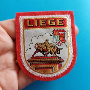 Op de afbeelding: Een rechthoekige patch met een rode rand en het woord "LIEGE" in gouden letters. De patch toont een stierenstandbeeld en een wapenschild. De achtergrond is wit en de patch wordt door een hand vastgehouden.