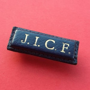 Op de afbeelding: Een donkerblauwe leren patch met gouden letters die "J.I.C.F." lezen.