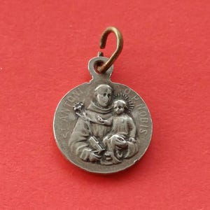 May include: A silver-colored religious pendant with a detailed relief of a saint holding a child. The pendant has a small loop at the top for hanging and is set against a red background.