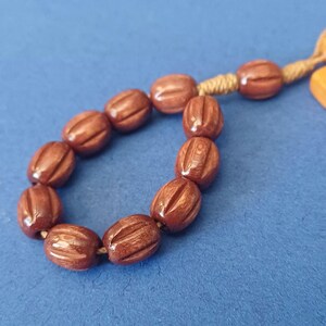 Pu&ograve; includere: Primo piano di una collana di perline marroni di forma ovale con linee verticali scolpite. Le perline sono infilate con un cordoncino beige, su uno sfondo blu.