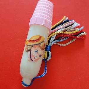 Pu&ograve; includere: Un ditale di plastica bianca con un top rosa e un fiocco colorato. Il ditale ha un'immagine di una ragazza con un cappello di paglia.
