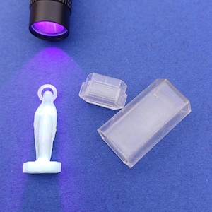 Puede incluir: Una linterna UV negra ilumina una pequeña figura blanca de la Virgen María y dos recipientes de plástico transparente sobre una superficie azul. La luz púrpura de la linterna resalta los detalles de la figura.