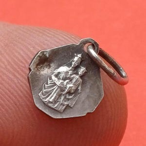 Op de afbeelding: Een kleine, zilverkleurige metalen hanger met een onregelmatige achthoekige vorm. Het oppervlak is voorzien van een verhoogd reliëf met twee figuren, één staand en één kleinere figuur ervoor, met een kleine ronde ring aan de bovenzijde.