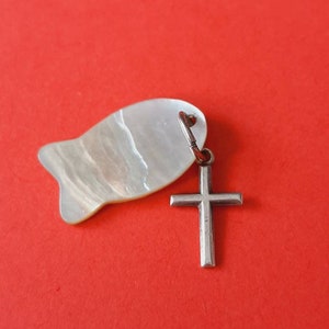 Op de afbeelding: Een hanger met een visvormige parelmoeren bedel en een zilveren kruis. De visbedel heeft een parelmoerglans en het kruis is een eenvoudig, klassiek ontwerp. De achtergrond is effen rood.