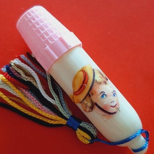 Pu&ograve; includere: Un ditale di plastica bianca con un tappo rosa e un fiocco colorato. Il ditale ha un adesivo di un bambino con un cappello di paglia.