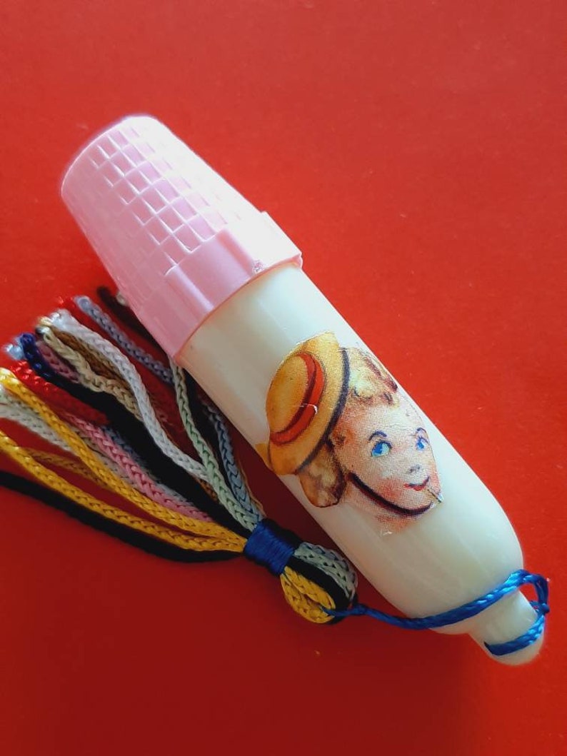 Pu&ograve; includere: Un ditale di plastica bianca con un tappo rosa e un fiocco colorato. Il ditale ha un adesivo di un bambino con un cappello di paglia.