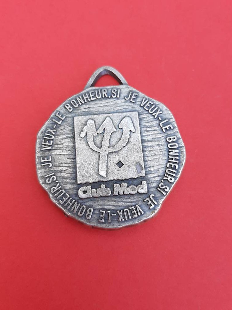 May include: A silver-toned metal keychain with a circular design. The keychain features a trident symbol and the text "IF VEUX-LE BONHEUR.SI JE VEUX-LE BONHEUR.SI JE VEUX-LE BONHEUR SI Clubs Med".