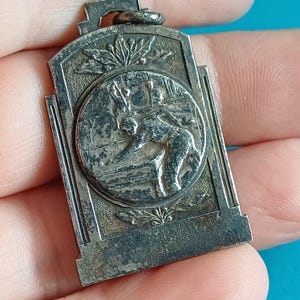 May include: A rectangular, silver-toned pendant with a detailed relief of a person playing a sport. The pendant has a circular central design with the figure in action, surrounded by decorative elements and a loop at the top for attachment.