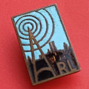 Puede incluir: Pin rectangular con fondo azul claro que representa una torre de radio y círculos concéntricos. El pin tiene un borde marrón oscuro y las letras ARL. Sobre un fondo rojo.