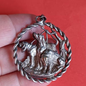 Può includere: Ciondolo in argento con un design dettagliato di tre cani da caccia in una cornice circolare con un bordo a corda.