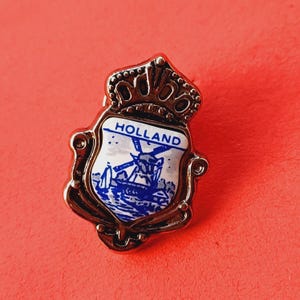 Puede incluir: Un pin de plata con un diseño de esmalte azul y blanco de un molino de viento y la palabra "HOLLAND". El pin tiene una corona en la parte superior.