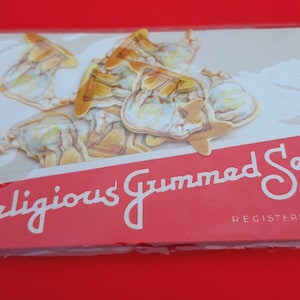 Peut inclure: Un paquet de timbres gommés religieux, une marque vintage de stickers représentant des images d'un animal blanc et brun, peut-être un kangourou, sur fond rouge. Le texte "Religious Gummed Seals" et "Registered" est imprimé sur l'emballage.