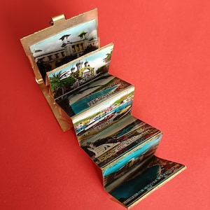 Pode incluir: Um suporte para postais em forma de acordeão de metal dourado, apresentando uma série de imagens impressas a cores de cidades costeiras italianas, incluindo San Remo e Bordighera, com etiquetas de texto a identificar cada local nos painéis.