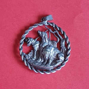 Puede incluir: Pendiente de plata con un diseño detallado de tres perros en un marco circular. El pendiente está hecho de plata y tiene un borde de cuerda.