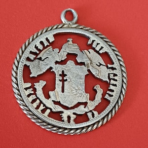 May include: A silver-colored pendant with a detailed design. The pendant features a central shield with a cross, surrounded by a cut-out pattern and a rope-like border. The background is a solid red.