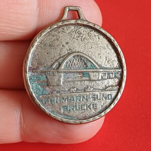 Puede incluir: Un colgante redondo plateado con un diseño de puente arqueado y el texto "Fehmarn-Sund Brücke". El colgante tiene un lazo en la parte superior para una cadena o cordón. El diseño del puente está detallado con sombreado cruzado.