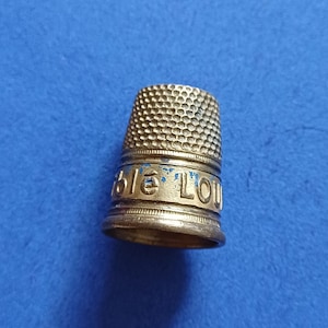 Peut inclure: Un dé à coudre en métal doré présentant un motif en nid d'abeille texturé sur le dôme supérieur et une bande lisse à la base. La bande affiche des lettres bleues partiellement effacées formant l'inscription "ble LOU" en relief.