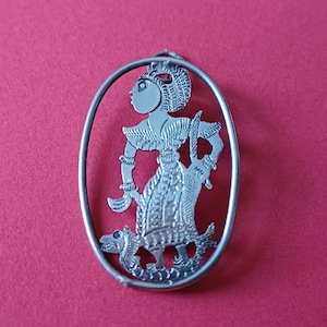 May include: A silver-colored pendant featuring a detailed cutout design of a figure in traditional attire, holding a weapon and standing on a creature. The pendant is set within an oval frame, with a loop at the top for a chain. The background is a solid red.
