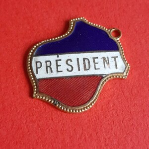 May include: A gold-colored metal pin with a blue, white, and red enamel design. The pin is shaped like a stylized map of a country and has the word "PR&Eacute;SIDENT" written on it.