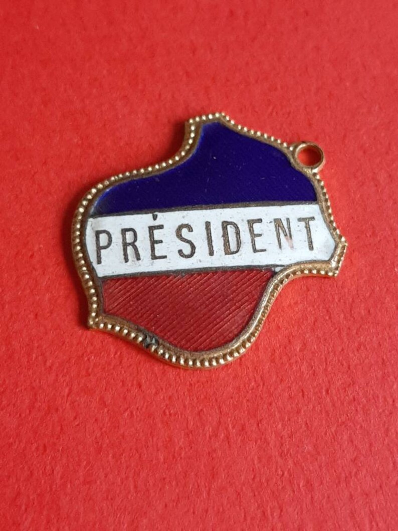May include: A gold-colored metal pin with a blue, white, and red enamel design. The pin is shaped like a stylized map of a country and has the word "PR&Eacute;SIDENT" written on it.