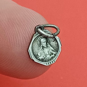 May include: A small, silver-toned, octagonal metal pendant featuring a raised relief image of a figure with a halo, possibly representing a religious icon. The pendant includes a circular jump ring attached to the top for hanging purposes.