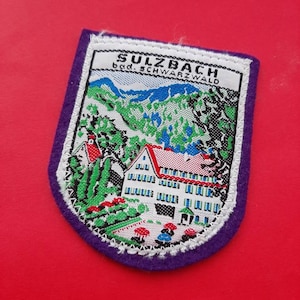 May include: A purple felt patch with a white embroidered image of a house and mountains. The text "Sulzbach bad. Schwarzwald" is embroidered in black.