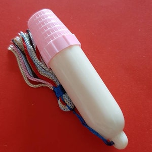 Pu&ograve; includere: Una coppetta mestruale in plastica bianca con un tappo rosa e un fiocco blu e bianco. La coppetta &egrave; su uno sfondo rosso.