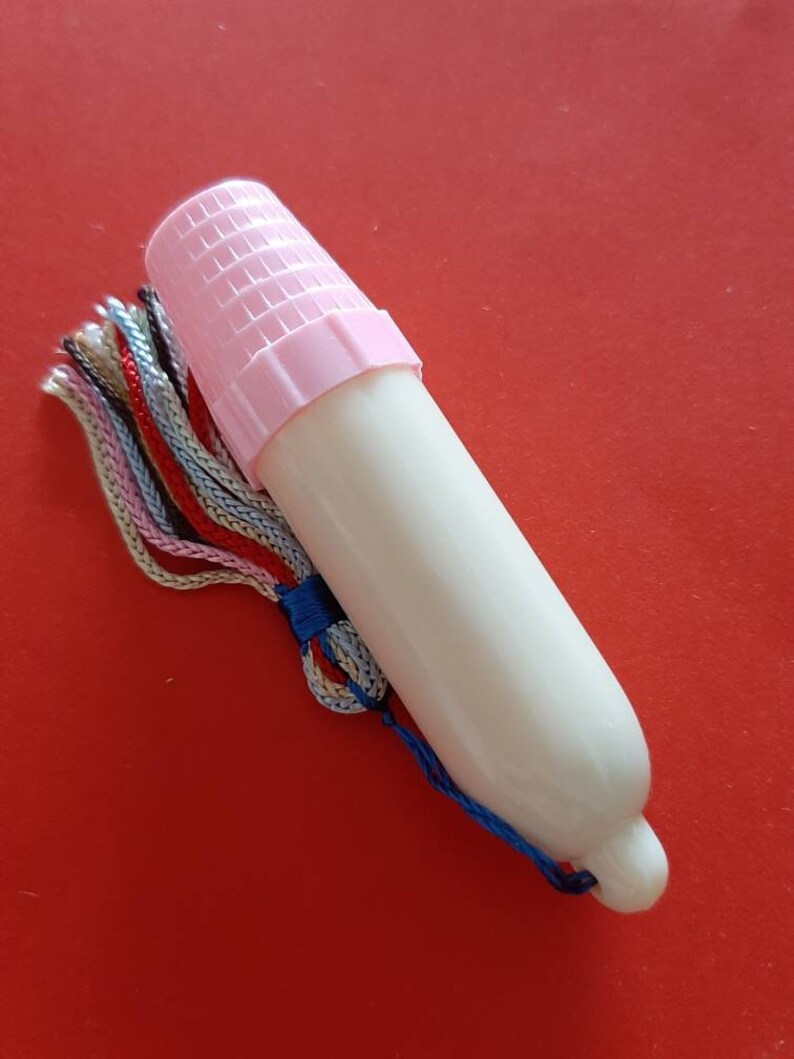 Pu&ograve; includere: Una coppetta mestruale in plastica bianca con un tappo rosa e un fiocco blu e bianco. La coppetta &egrave; su uno sfondo rosso.