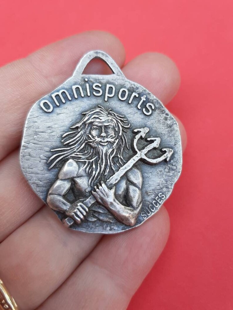 May include: Silver-toned metal keychain with a raised image of a man holding a trident. The keychain is round and has the words "omnisports" and "succes" inscribed on it.