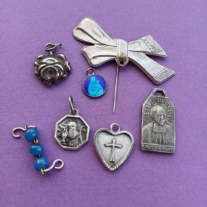 Puede incluir: Una colección de dijes y colgantes de plata, que incluyen una rosa, un corazón con una cruz, una figura religiosa, un dije de esmalte azul y un lazo de plata.