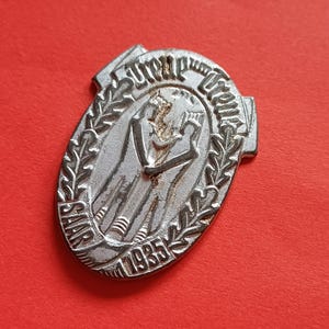 May include: A silver-colored oval-shaped pin with the text "SAAR 1935" and an image of two figures. The pin is surrounded by a laurel wreath and the text "Unser Treue". The pin is on a red background.