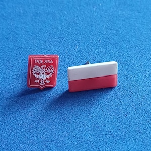 Può includere: Due spille della bandiera polacca. Una spilla è uno scudo rosso con un'aquila bianca e la scritta "POLSKA". L'altra spilla è di forma rettangolare con la parte superiore bianca e inferiore rossa. Entrambe le spille sono su una superficie blu.
