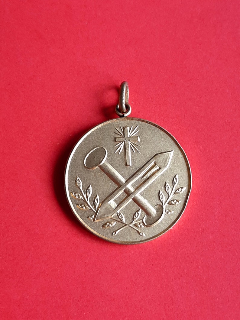 May include: A gold-toned pendant with a cross and two nails, surrounded by a wreath of leaves.