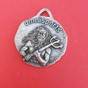 May include: Silver-toned metal keychain with a raised image of a man holding a trident. The keychain is round and has the words "omnisports" and "succes" inscribed on it.