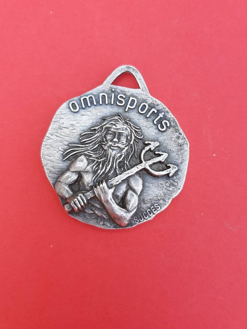 May include: Silver-toned metal keychain with a raised image of a man holding a trident. The keychain is round and has the words "omnisports" and "succes" inscribed on it.