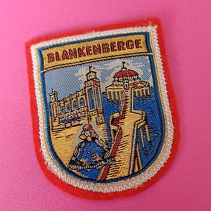 Op de afbeelding: Een rechthoekige patch met het woord "BLANKENBERGE" bovenaan. De patch toont een gedetailleerde illustratie van gebouwen en een figuur in een blauwe outfit. De patch heeft een rode rand en staat op een roze achtergrond.