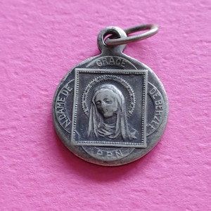 Op de afbeelding: Zilveren hanger met een religieus beeld van een vrouw in een sluier, omringd door een halo. De hanger is ovaalvormig en heeft de woorden "GRACE DE" en "MADAME DE" boven en onder, respectievelijk. De hanger heeft ook de letters "P.P.N." onderaan.