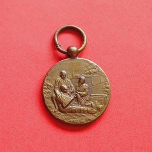 Op de afbeelding: Een bronzen cirkelvormige hanger met een gedetailleerde reliëfscène van drie figuren. De hanger heeft een lus aan de bovenkant om op te hangen. De achtergrond is effen rood.