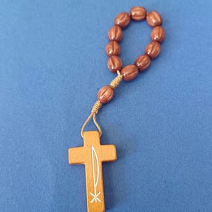 Pu&ograve; includere: Un rosario in legno con perline ovali marroni e un ciondolo a forma di croce in legno. La croce presenta un disegno bianco. Il rosario &egrave; su sfondo blu.