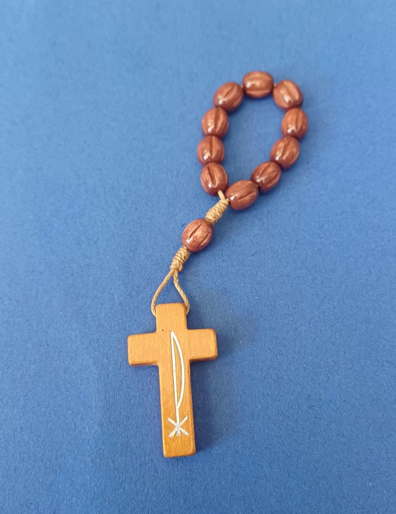 Pu&ograve; includere: Un rosario in legno con perline ovali marroni e un ciondolo a forma di croce in legno. La croce presenta un disegno bianco. Il rosario &egrave; su sfondo blu.